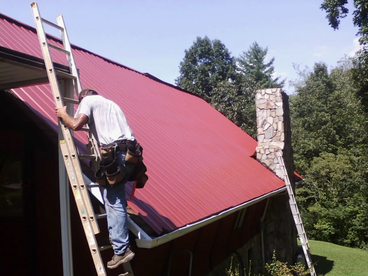 Roofer on a ladder beside a red metal roof and stone chimney — Soffit & Fascia Repair by Roofing Brothers in Buena Park